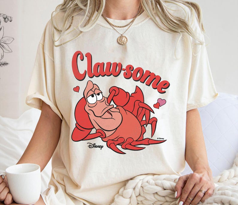 May include: Cream-colored t-shirt featuring a cartoon illustration of Sebastian the crab from Disney's The Little Mermaid. The text "Claw-some" is written in red above the crab. Two small hearts are also included.