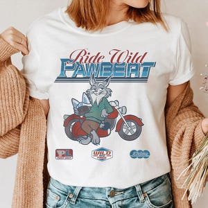 Puede incluir: Camiseta blanca con un conejo de dibujos animados montado en una moto roja. El texto "Ride Wild Pawbert" est&aacute; en azul y rojo. El conejo lleva un su&eacute;ter verde y pantalones marrones. Gr&aacute;ficos adicionales debajo.