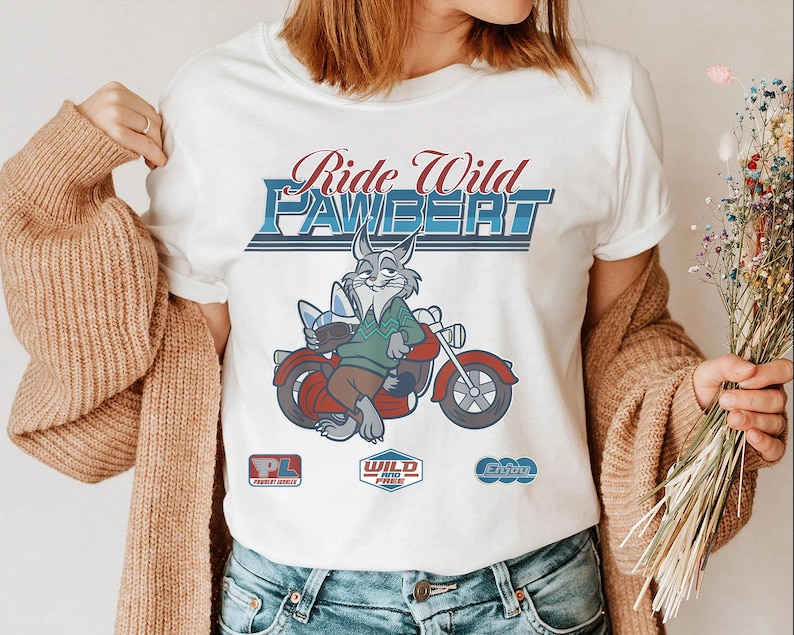 Puede incluir: Camiseta blanca con un conejo de dibujos animados montado en una moto roja. El texto "Ride Wild Pawbert" est&aacute; en azul y rojo. El conejo lleva un su&eacute;ter verde y pantalones marrones. Gr&aacute;ficos adicionales debajo.