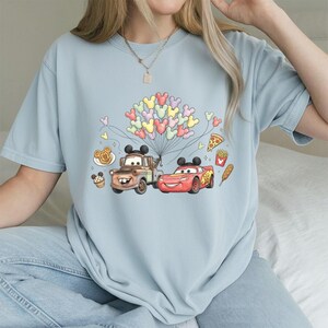 May include: Pale blue t-shirt with a Cars movie graphic featuring Mater and Lightning McQueen, holding Mickey Mouse balloons. The design includes food icons like pizza and fries. The shirt is made of a soft material.