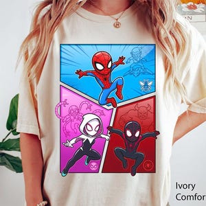 May include: Ivory Comfort Color t-shirt featuring a comic book style graphic of Spider-Man and other characters. The graphic is divided into panels with a blue, pink, and red background. The text "Ivory Comfort Color" is printed on the bottom right.
