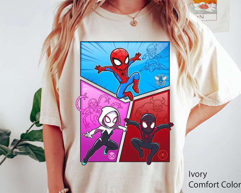 May include: Ivory Comfort Color t-shirt featuring a comic book style graphic of Spider-Man and other characters. The graphic is divided into panels with a blue, pink, and red background. The text "Ivory Comfort Color" is printed on the bottom right.
