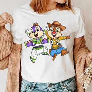 May include: White t-shirt featuring cartoon characters Chip and Dale dressed as Buzz Lightyear and Woody. Chip is in a Buzz Lightyear costume with a purple and green color scheme. Dale is in a Woody costume with a cowboy hat and blue jeans.