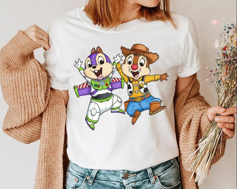 May include: White t-shirt featuring cartoon characters Chip and Dale dressed as Buzz Lightyear and Woody. Chip is in a Buzz Lightyear costume with a purple and green color scheme. Dale is in a Woody costume with a cowboy hat and blue jeans.