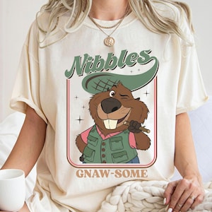 May include: Cream-colored t-shirt featuring a cartoon beaver winking and holding a stick. The beaver wears a green vest and a hat. The text "Nibbles" in green and "GNAW-SOME" in brown are above and below the beaver illustration.