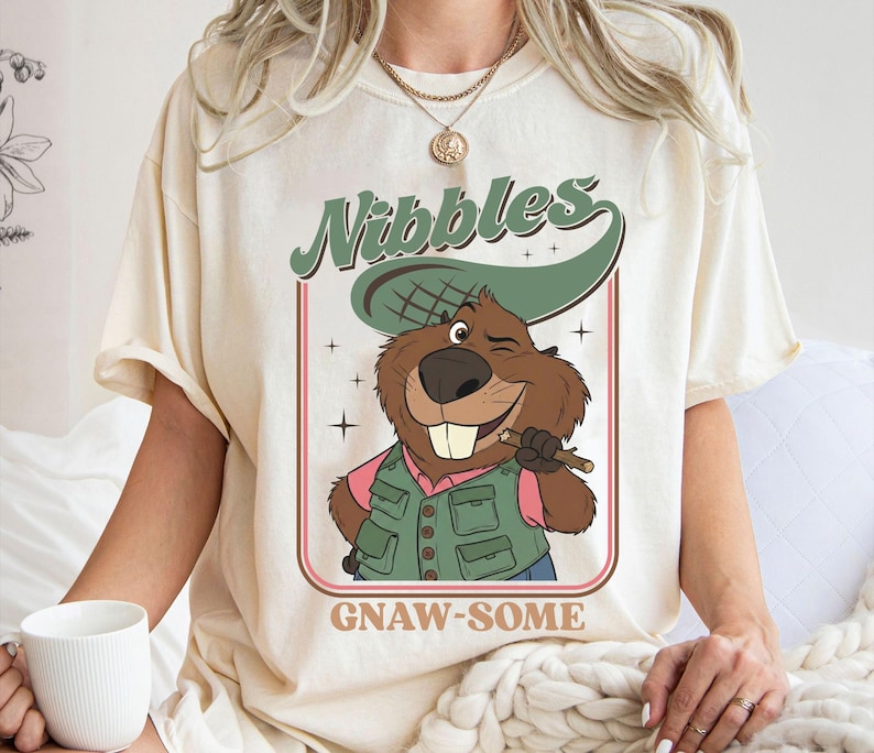 May include: Cream-colored t-shirt featuring a cartoon beaver winking and holding a stick. The beaver wears a green vest and a hat. The text "Nibbles" in green and "GNAW-SOME" in brown are above and below the beaver illustration.