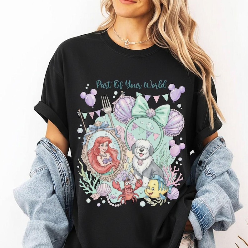 May include: A black t-shirt featuring a graphic print with the text Part Of Your World, an illustration of a mermaid, a dog, a crab, a fish, a fork, a large bow, and purple balloons, all arranged in a colorful underwater-themed design.