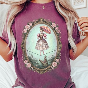 May include: A purple t-shirt featuring a framed illustration of a woman balancing on a tightrope above a swamp with an alligator. The woman holds a pink parasol. The frame is decorated with roses.