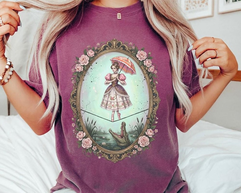 May include: A purple t-shirt featuring a framed illustration of a woman balancing on a tightrope above a swamp with an alligator. The woman holds a pink parasol. The frame is decorated with roses.