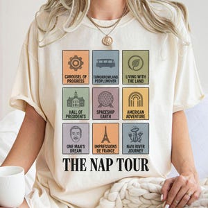 May include: Cream-colored t-shirt featuring a grid of colorful squares with theme park attraction icons and names. The text "THE NAP TOUR" is printed below the squares. The shirt is made of soft material.