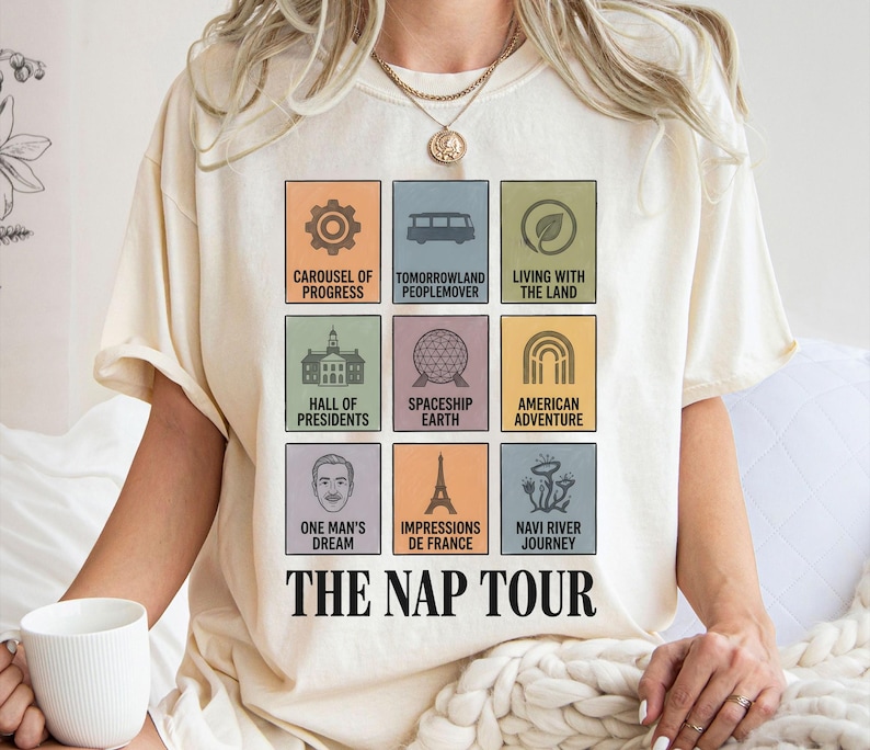 May include: Cream-colored t-shirt featuring a grid of colorful squares with theme park attraction icons and names. The text "THE NAP TOUR" is printed below the squares. The shirt is made of soft material.