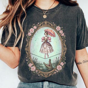 May include: Dark gray t-shirt featuring a framed illustration of a woman with an umbrella, balancing on a tightrope above a lake with an alligator. The frame is adorned with pink roses. The woman is wearing a dress with pink and brown accents.
