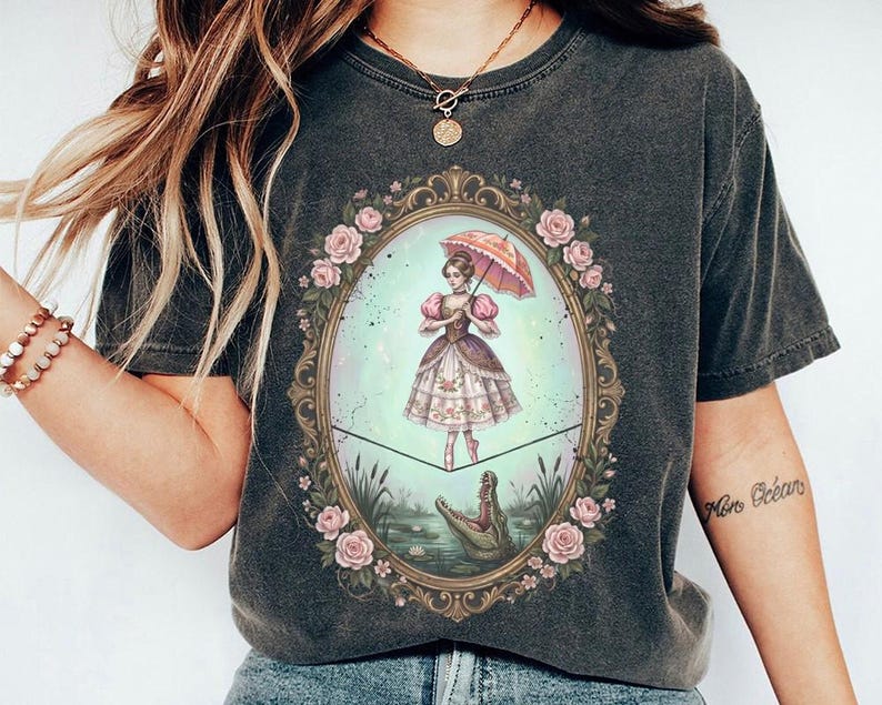 May include: Dark gray t-shirt featuring a framed illustration of a woman with an umbrella, balancing on a tightrope above a lake with an alligator. The frame is adorned with pink roses. The woman is wearing a dress with pink and brown accents.