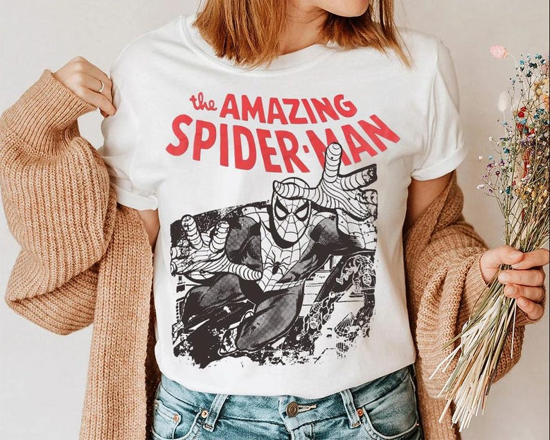 May include: A white short-sleeved t-shirt featuring a graphic print of the comic book character Spider-Man in a dynamic pose. Above the illustration, the text reads "the AMAZING SPIDER-MAN" in bold red lettering. The design includes black and white ink details.