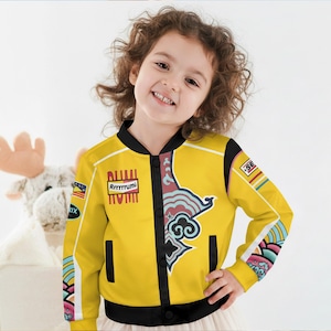 May include: A yellow bomber jacket with black trim and colorful graphic designs. The jacket features the text "Rrrrrrumi" and other decorative elements. The sleeves have additional patterns in shades of blue, pink, and white. The jacket is worn by a child.