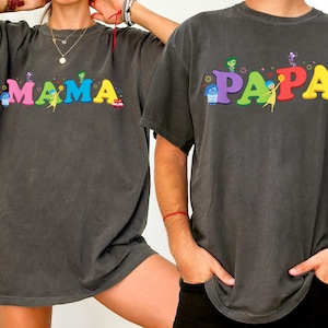 May include: Two charcoal gray t-shirts with the words "MAMA" and "PAPA" in colorful, cartoon-style lettering. The shirts feature characters from the animated movie Inside Out. The shirts are short-sleeved.