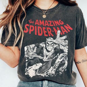 May include: A charcoal gray short-sleeve t-shirt featuring a graphic print of the comic book character Spider-Man in a dynamic pose. The text "the AMAZING SPIDER-MAN" is printed in bold red letters above the black and white illustration.