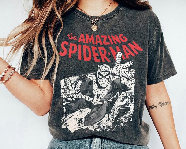 May include: A charcoal gray short-sleeve t-shirt featuring a graphic print of the comic book character Spider-Man in a dynamic pose. The text "the AMAZING SPIDER-MAN" is printed in bold red letters above the black and white illustration.
