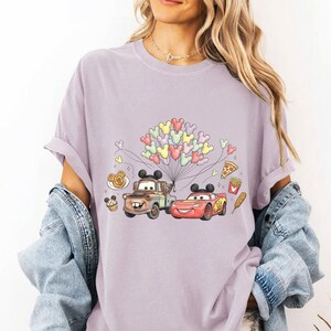 May include: A lilac t-shirt with a graphic of Mater and Lightning McQueen from the film Cars, holding Mickey Mouse-shaped balloons. The design includes food icons and Mickey Mouse icons.