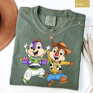 May include: Moss green t-shirt featuring a graphic of two cartoon characters, one dressed as Buzz Lightyear and the other as Woody. The shirt has a crew neck and short sleeves. A gold necklace with a pendant is draped across the shirt.
