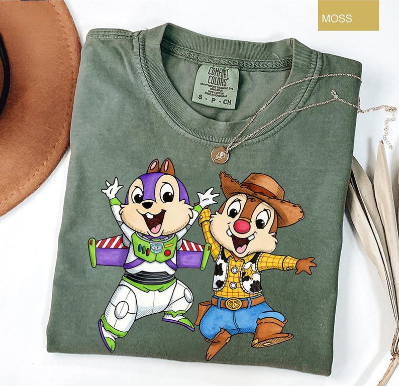 May include: Moss green t-shirt featuring a graphic of two cartoon characters, one dressed as Buzz Lightyear and the other as Woody. The shirt has a crew neck and short sleeves. A gold necklace with a pendant is draped across the shirt.