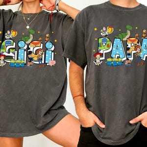 K&ouml;nnte beinhalten: Zwei anthrazitfarbene T-Shirts mit den W&ouml;rtern "GIGI" und "PAPA" in einer bunten Schriftart im Cartoon-Stil. Die Shirts zeigen Charaktere aus einem beliebten Animationsfilm, darunter ein Dinosaurier, ein Cowboy und ein Weltraumranger.