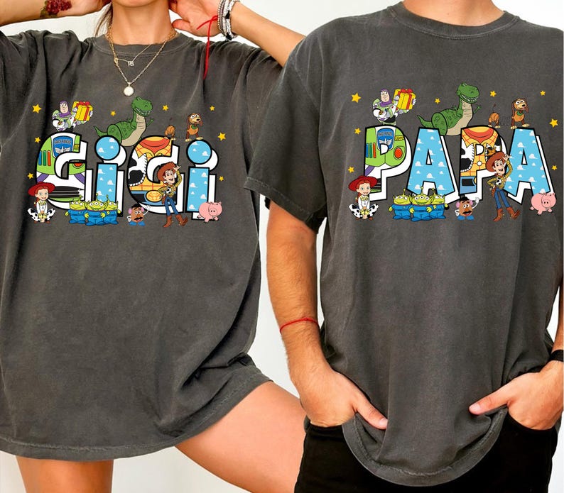 K&ouml;nnte beinhalten: Zwei anthrazitfarbene T-Shirts mit den W&ouml;rtern "GIGI" und "PAPA" in einer bunten Schriftart im Cartoon-Stil. Die Shirts zeigen Charaktere aus einem beliebten Animationsfilm, darunter ein Dinosaurier, ein Cowboy und ein Weltraumranger.