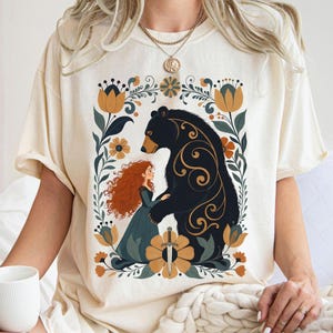 Op de afbeelding: Cr&egrave;mekleurig T-shirt met een afbeelding van een vrouw met rood haar en een beer, omringd door bloemmotieven. De beer is zwart met gouden accenten. De vrouw draagt een tealkleurige jurk. Een zwaard staat onderaan het ontwerp.
