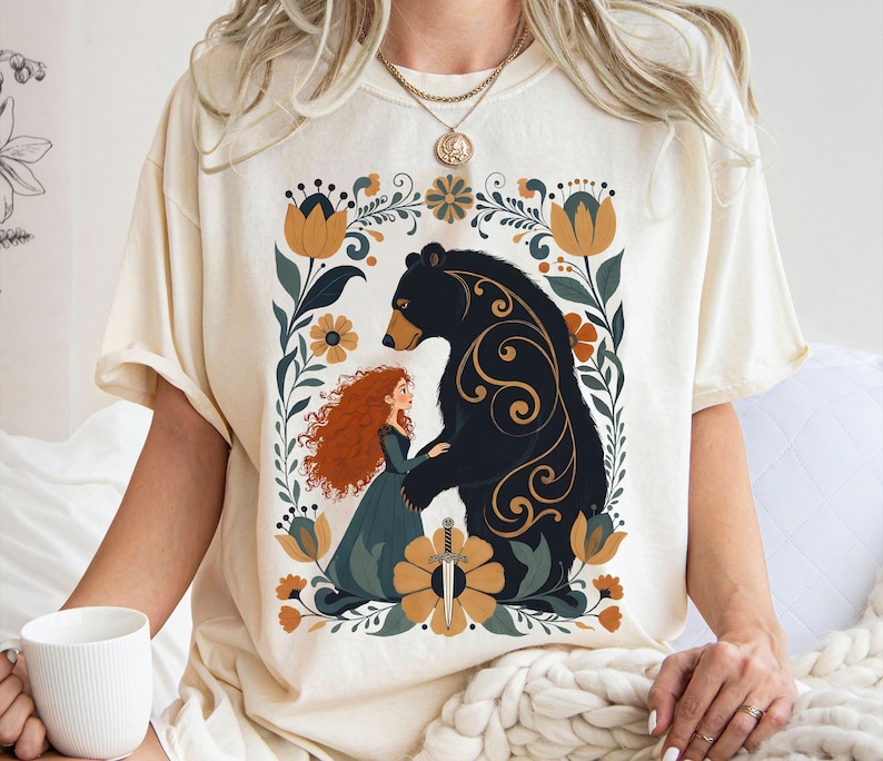 Op de afbeelding: Cr&egrave;mekleurig T-shirt met een afbeelding van een vrouw met rood haar en een beer, omringd door bloemmotieven. De beer is zwart met gouden accenten. De vrouw draagt een tealkleurige jurk. Een zwaard staat onderaan het ontwerp.