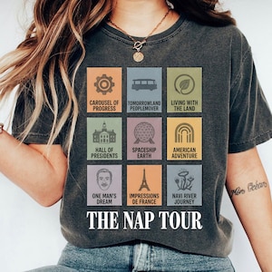 May include: Dark gray t-shirt featuring colorful squares with theme park attraction icons and text. The shirt reads "THE NAP TOUR" in white font. The shirt has a vintage, worn look.