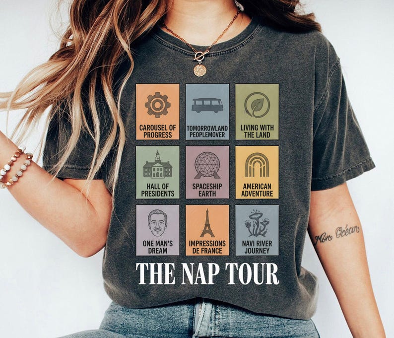 May include: Dark gray t-shirt featuring colorful squares with theme park attraction icons and text. The shirt reads "THE NAP TOUR" in white font. The shirt has a vintage, worn look.