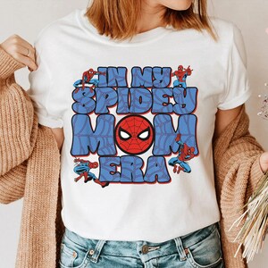 May include: White t-shirt featuring a graphic design with the text "IN MY SPIDEY MOM ERA" in blue bubble letters. The design includes a red Spider-Man mask and Spider-Man figures. A casual, fun, and colorful design.