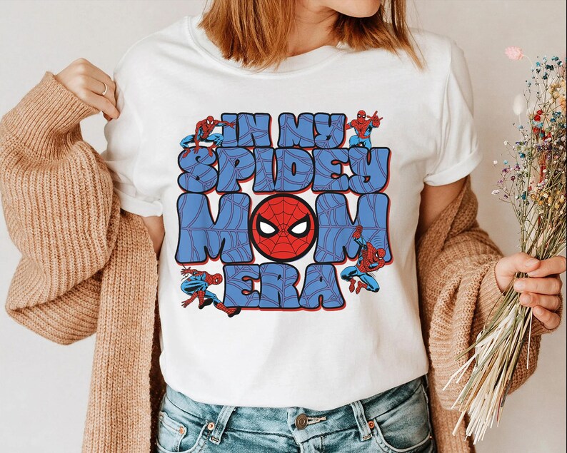 May include: White t-shirt featuring a graphic design with the text "IN MY SPIDEY MOM ERA" in blue bubble letters. The design includes a red Spider-Man mask and Spider-Man figures. A casual, fun, and colorful design.