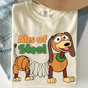 K&ouml;nnte beinhalten: Cremefarbenes T-Shirt mit einem Cartoon-Hund und dem Text "Abs of Steel". Der Hund ist braun und beige mit einem federartigen K&ouml;rper. Das T-Shirt hat einen Rundhalsausschnitt und kurze &Auml;rmel. Der Text ist orange und gr&uuml;n.