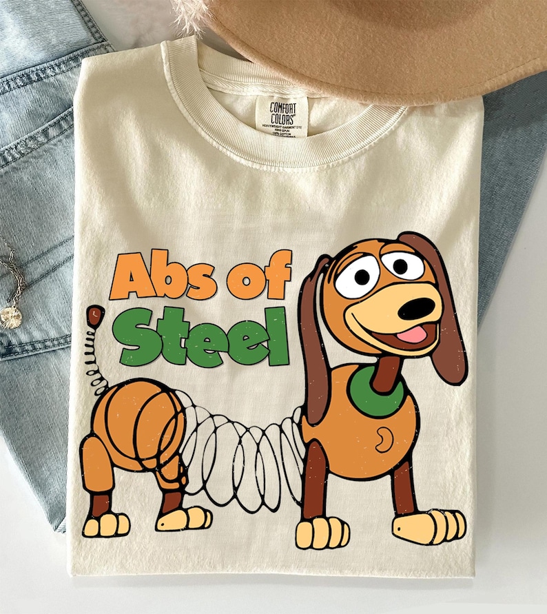 K&ouml;nnte beinhalten: Cremefarbenes T-Shirt mit einem Cartoon-Hund und dem Text "Abs of Steel". Der Hund ist braun und beige mit einem federartigen K&ouml;rper. Das T-Shirt hat einen Rundhalsausschnitt und kurze &Auml;rmel. Der Text ist orange und gr&uuml;n.