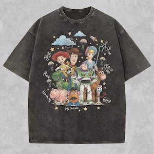May include: A charcoal gray short-sleeve t-shirt featuring a graphic print of Toy Story characters including Woody, Buzz Lightyear, Jessie, Rex, Hamm, Mr. Potato Head, Bo Peep, Bullseye, and Forky, surrounded by stars, clouds, and handwritten names.