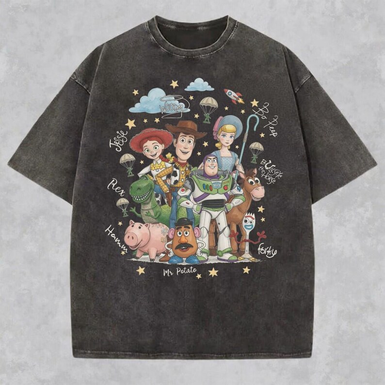 May include: A charcoal gray short-sleeve t-shirt featuring a graphic print of Toy Story characters including Woody, Buzz Lightyear, Jessie, Rex, Hamm, Mr. Potato Head, Bo Peep, Bullseye, and Forky, surrounded by stars, clouds, and handwritten names.