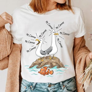May include: White t-shirt featuring a cartoon illustration of two seagulls on a rock, with the text "Mr. Connor" emanating from them. A cartoon clownfish is swimming below. The design is in black, white, orange, and blue.