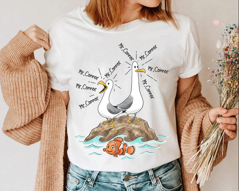 May include: White t-shirt featuring a cartoon illustration of two seagulls on a rock, with the text "Mr. Connor" emanating from them. A cartoon clownfish is swimming below. The design is in black, white, orange, and blue.