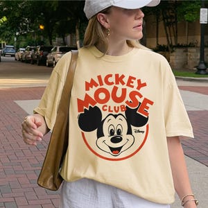 May include: Cream-colored t-shirt featuring a Mickey Mouse graphic. The design includes the words "Mickey Mouse Club" in red, with Mickey's face in the center. The Disney logo is at the bottom right.