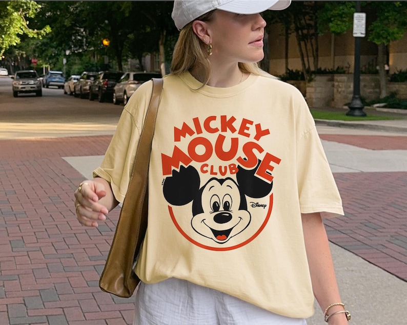 May include: Cream-colored t-shirt featuring a Mickey Mouse graphic. The design includes the words "Mickey Mouse Club" in red, with Mickey's face in the center. The Disney logo is at the bottom right.