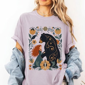 Op de afbeelding: Lichtpaarse t-shirt met een afbeelding van een roodharige vrouw en een zwarte beer, omringd door bloemen. De vrouw draagt een groene jurk en houdt een zwaard vast. De beer heeft gouden krullen.