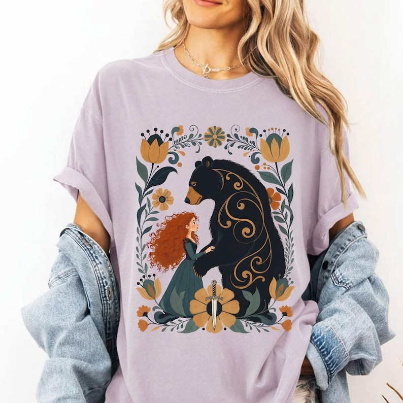 Op de afbeelding: Lichtpaarse t-shirt met een afbeelding van een roodharige vrouw en een zwarte beer, omringd door bloemen. De vrouw draagt een groene jurk en houdt een zwaard vast. De beer heeft gouden krullen.