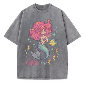 May include: A charcoal grey acid-wash t-shirt featuring a graphic print of Ariel from The Little Mermaid with pink hair, a green tail, and a yellow fish friend. The design includes small seahorses and bubbles, with the text The Little Mermaid.