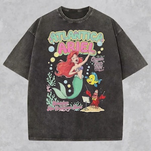 May include: A charcoal gray short-sleeve t-shirt featuring a graphic print of Ariel, Flounder, and Sebastian. The text reads Atlantica Ariel Ocean Tour 1991 and Sebastian with the Hot Crustacean Band. The design includes colorful coral and bubbles.