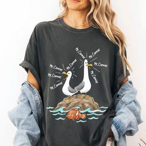 May include: Dark gray t-shirt featuring a cartoon design of two seagulls on a rock, with the text "Mr. Connor" emanating from them. Below, an orange and white clownfish swims in blue water. A fun, whimsical design.