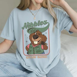 May include: Light blue t-shirt featuring a cartoon beaver winking and holding a stick. The beaver wears a green vest and hat, with the word "Nibbles" above and "Gnaw-some" below in a retro design.