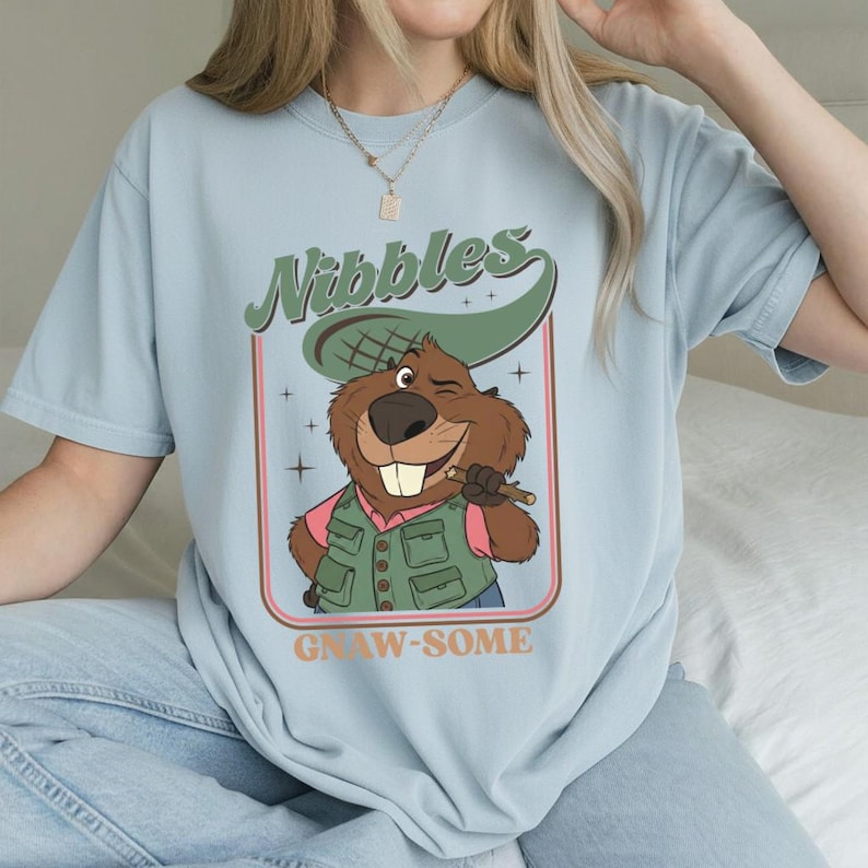 May include: Light blue t-shirt featuring a cartoon beaver winking and holding a stick. The beaver wears a green vest and hat, with the word "Nibbles" above and "Gnaw-some" below in a retro design.