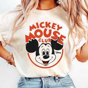 May include: Cream-colored t-shirt featuring a red and black Mickey Mouse Club graphic. The design includes Mickey Mouse's face within a red circle, with the words "Mickey Mouse Club" in red. The Disney logo is also visible.