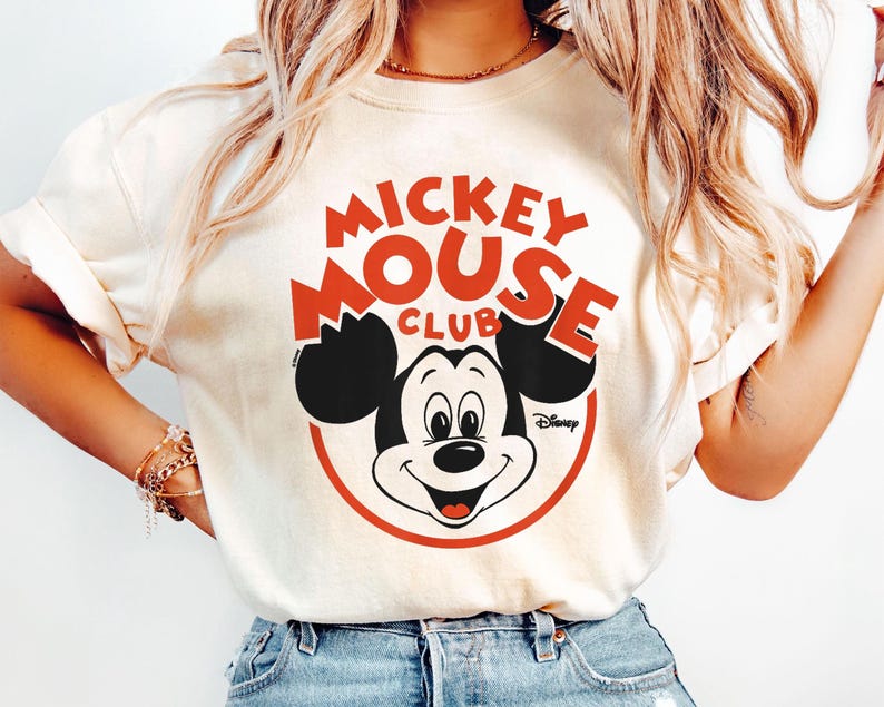 May include: Cream-colored t-shirt featuring a red and black Mickey Mouse Club graphic. The design includes Mickey Mouse's face within a red circle, with the words "Mickey Mouse Club" in red. The Disney logo is also visible.